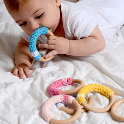 Baby holding wooden teething ring with blue crochet cotton to mouth with dark pink, light pink and yellow rings lying on blanket